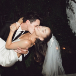 Ariana Grande in a white gown and veil being carried in a bridal position by husband Dalton Gomez, who is in a black suit with a white shirt and black tie.