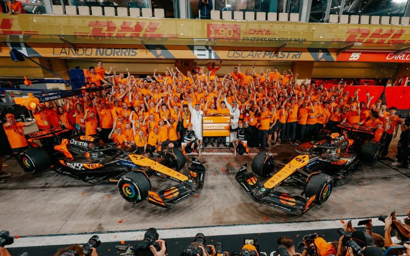 A group overhead picture of McLaren F1 team garage, team principal Andrea Stella, Owner Zac Brown, and drivers Lando Norris and Oscar Piastri celebrating Norris' win in Abu Dhabi and the team's constructors' championship title win.